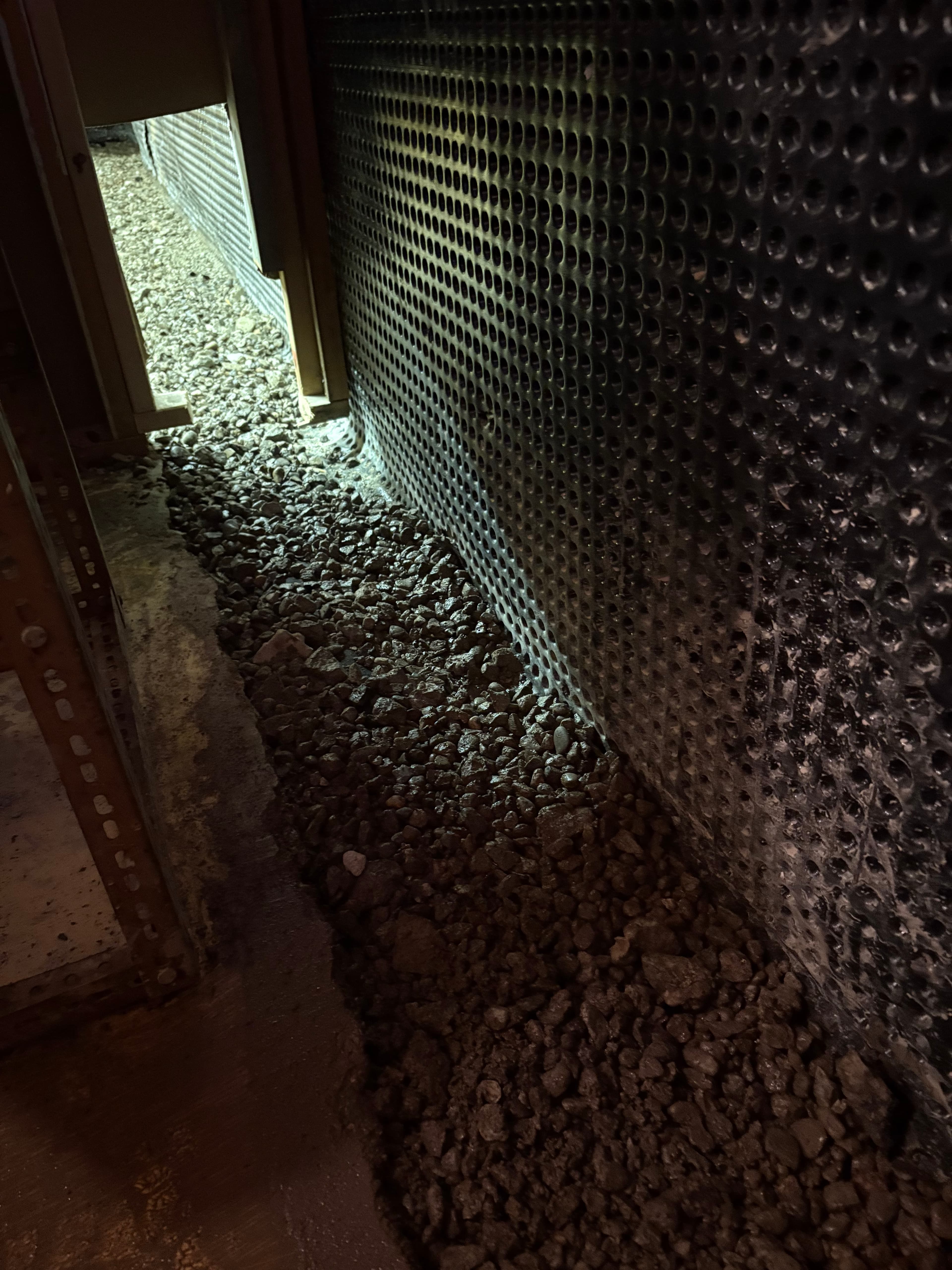 Dimpled foundation drainage membrane next to a trench of crushed gravel in a dark space.